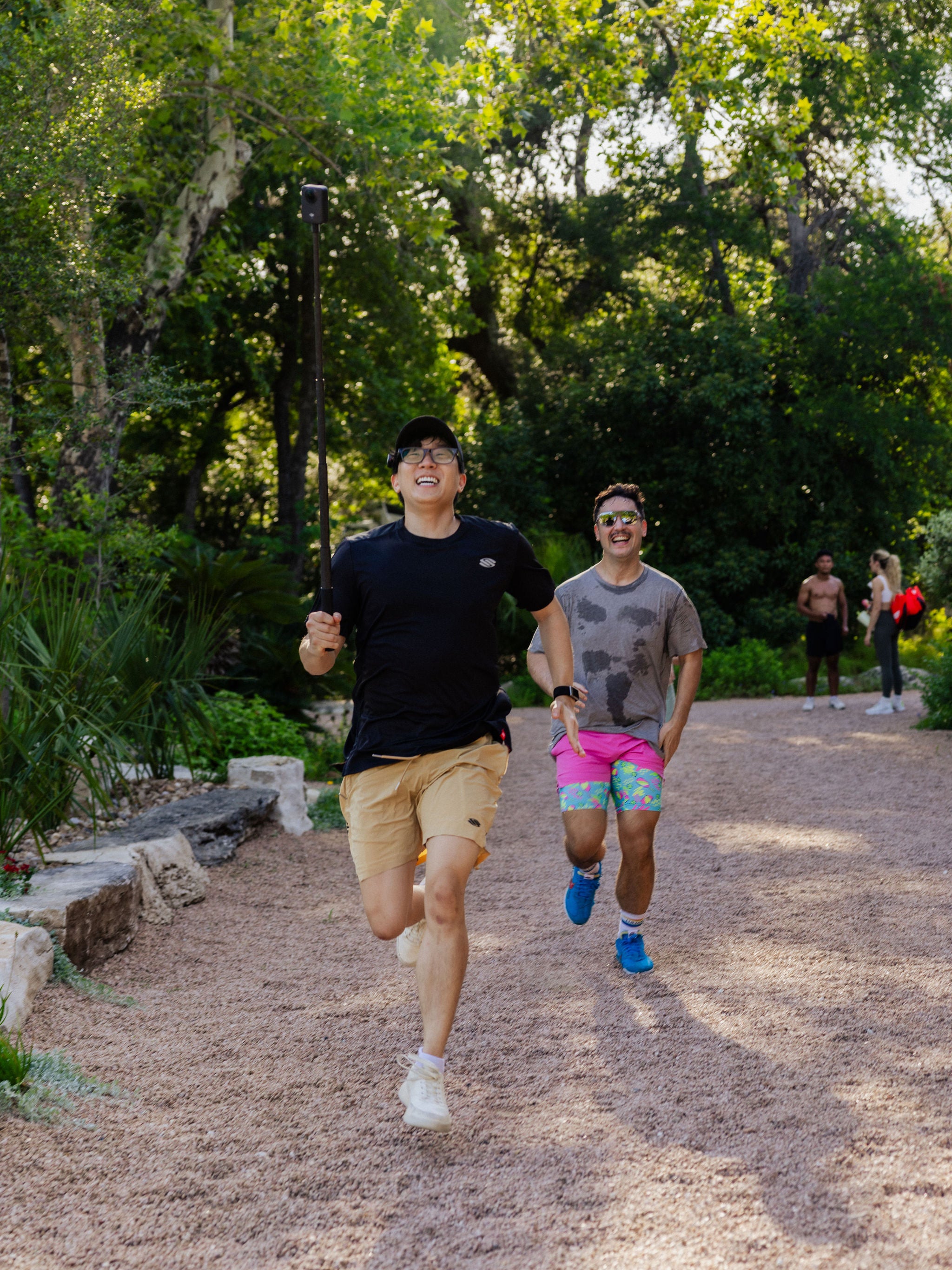 Two men run and smile as the man in the lead holds a video camera to record them. 