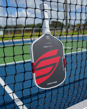 Black Selkirk LABS Boomstik pickleball paddle hanging on the net of an outdoor court
