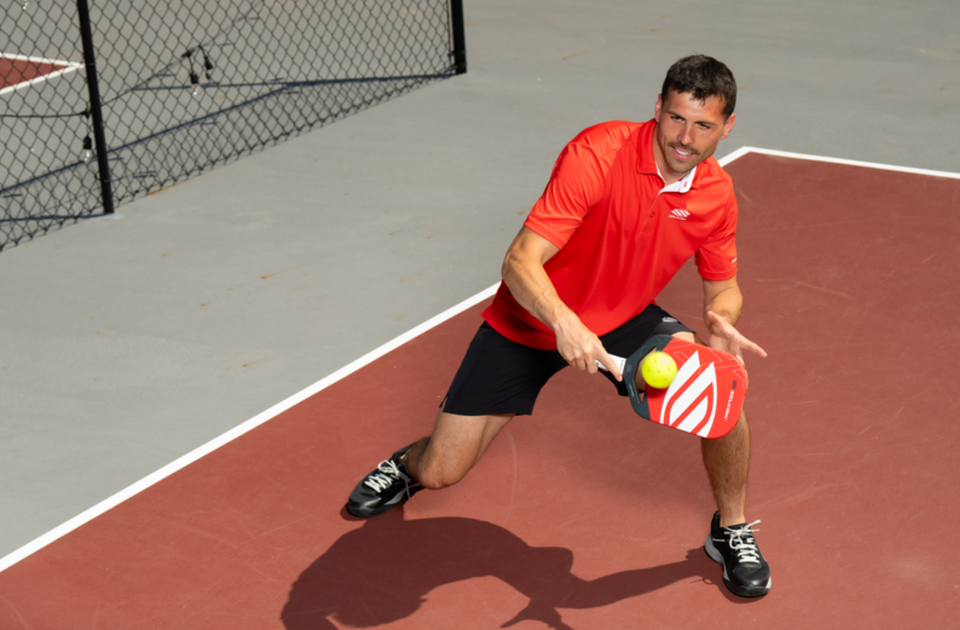 Man in red Selkirk shirt kneeling on a pickleball court reaching to strike a ball with a red Selkirk paddle