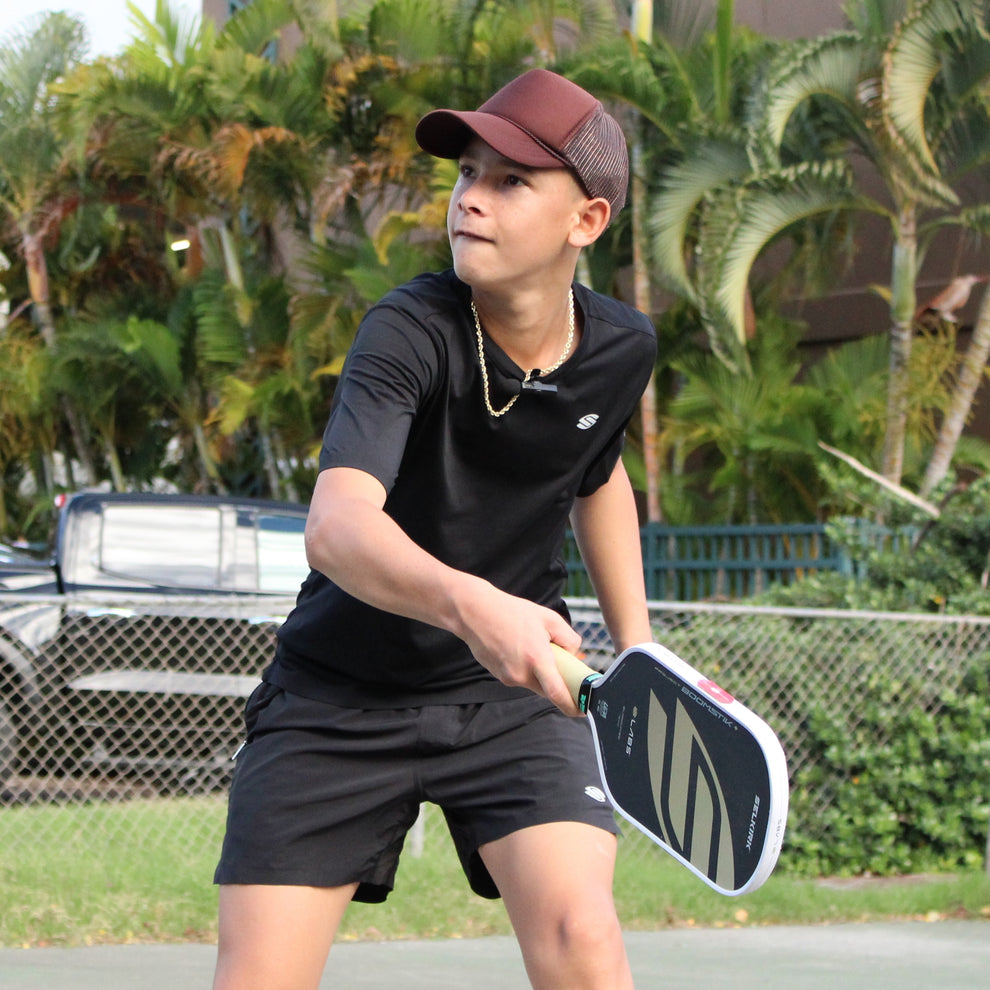 Teenage boy in a brown cap and black shirt reaching with a pickleball paddle on an outdoor court