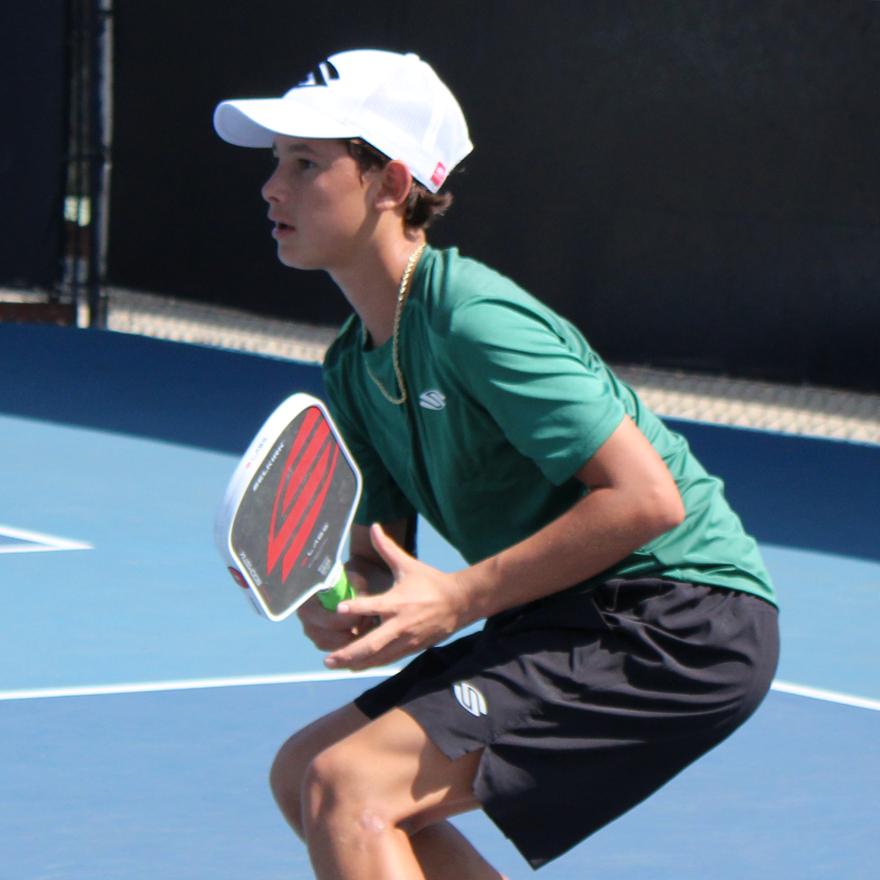 Player crouched ready to hit a pickleball with a paddle on an outdoor blue court, wearing green shirt and white cap
