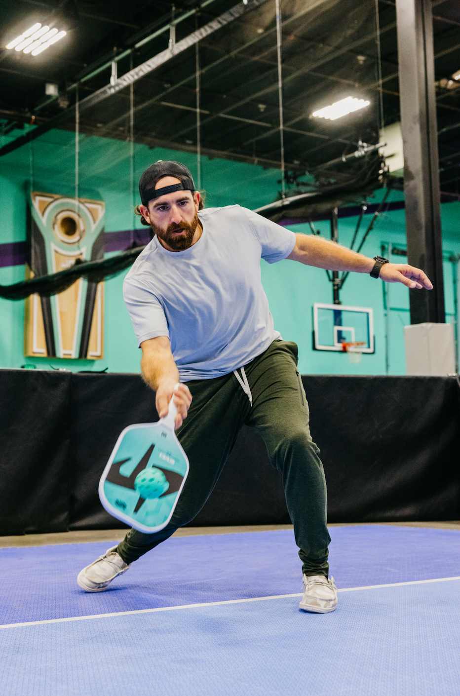 A man in a gray shirt and green pants playing pickleball with the SLK by Selkirk paddle on a blue court.