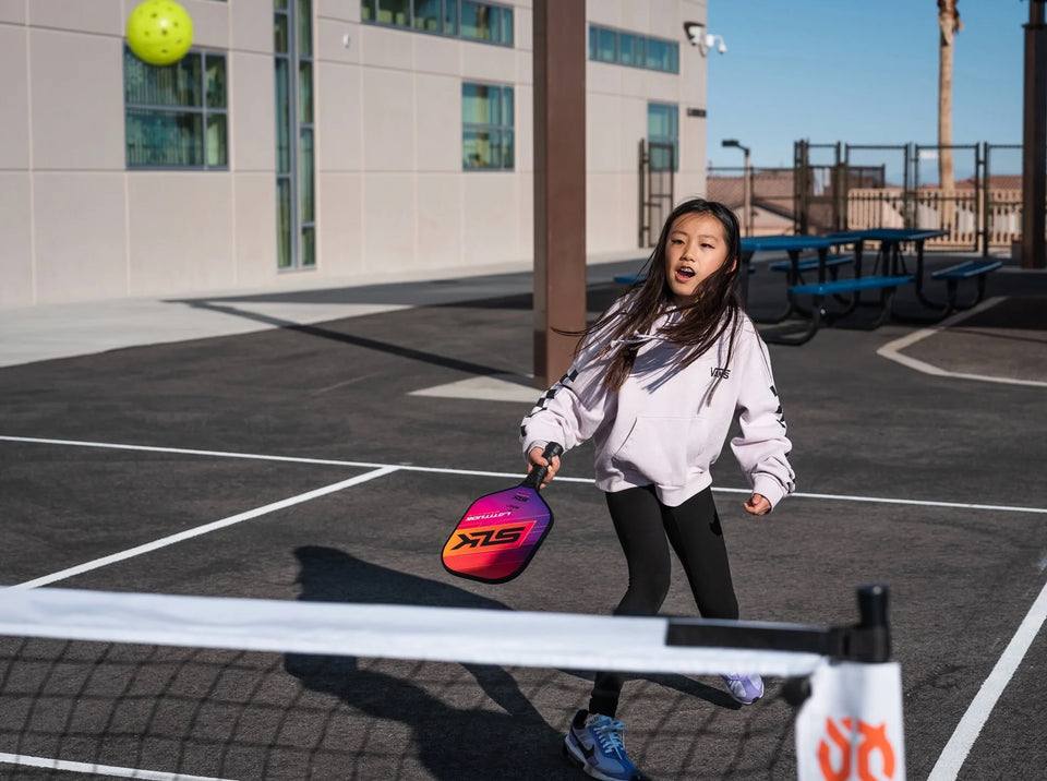 Young girl in a VANS hoodie playing pickleball on an outdoor court, swinging a colorful paddle toward a lime ball.