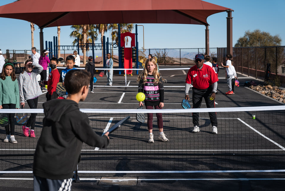 Children and instructor playing pickleball on outdoor courts under shade; girl wearing a sweater reading 'ON THE NAUGHTY LIST'.