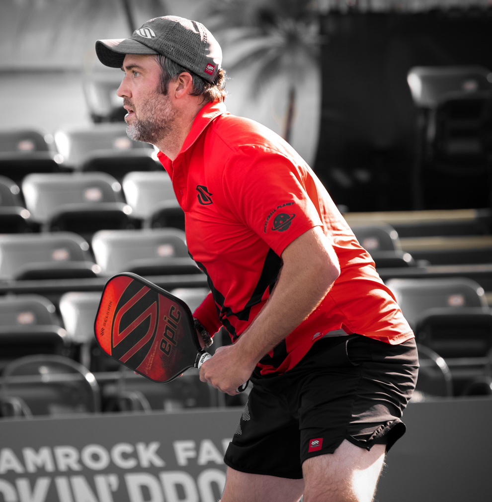 Man in red shirt poised to hit a pickleball, holding a paddle labeled 'epic', with stadium seating behind him.