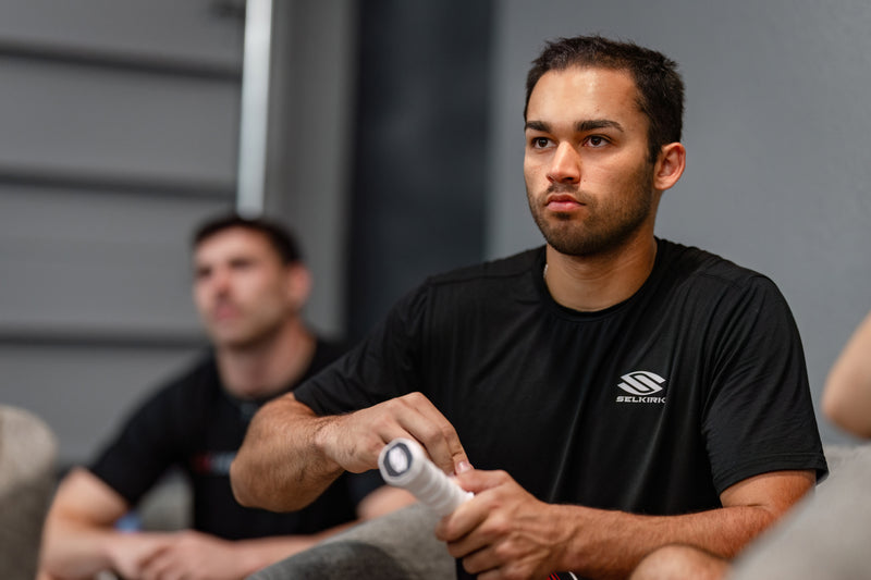 Man in a black shirt with a selkirk logo sitting on a couch, holding a pickleball paddle