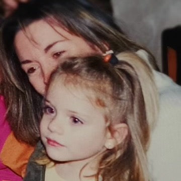 A woman with long hair and a young girl with a ponytail, both smiling together.