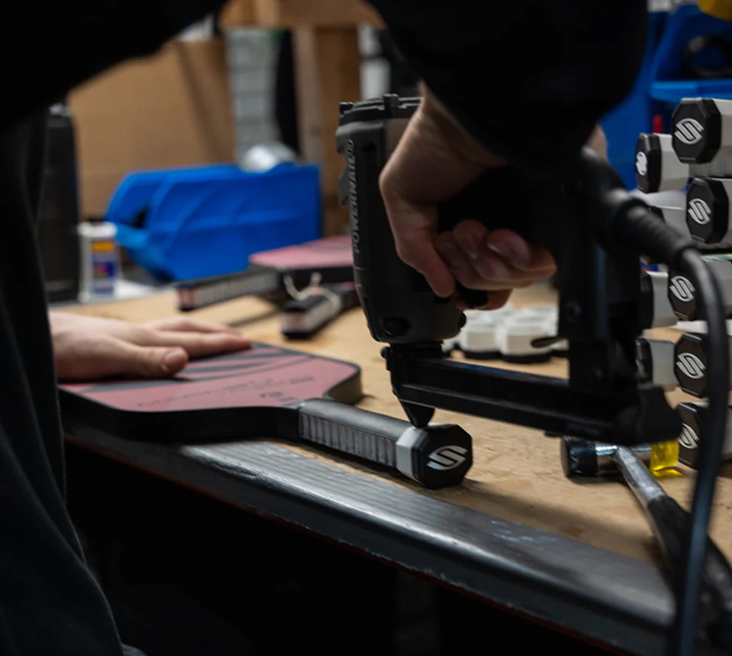 Close-up of a Selkirk Sport technician assembling a pickleball paddle by securing the handle with precision tools in a workshop, showcasing detailed craftsmanship and branded components.