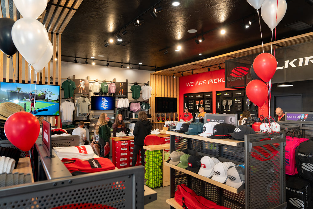 Retail pickleball store interior with merchandise tables, hats and paddles, red 'WE ARE PICKL' wall sign and SELKIRK and LUXX branding