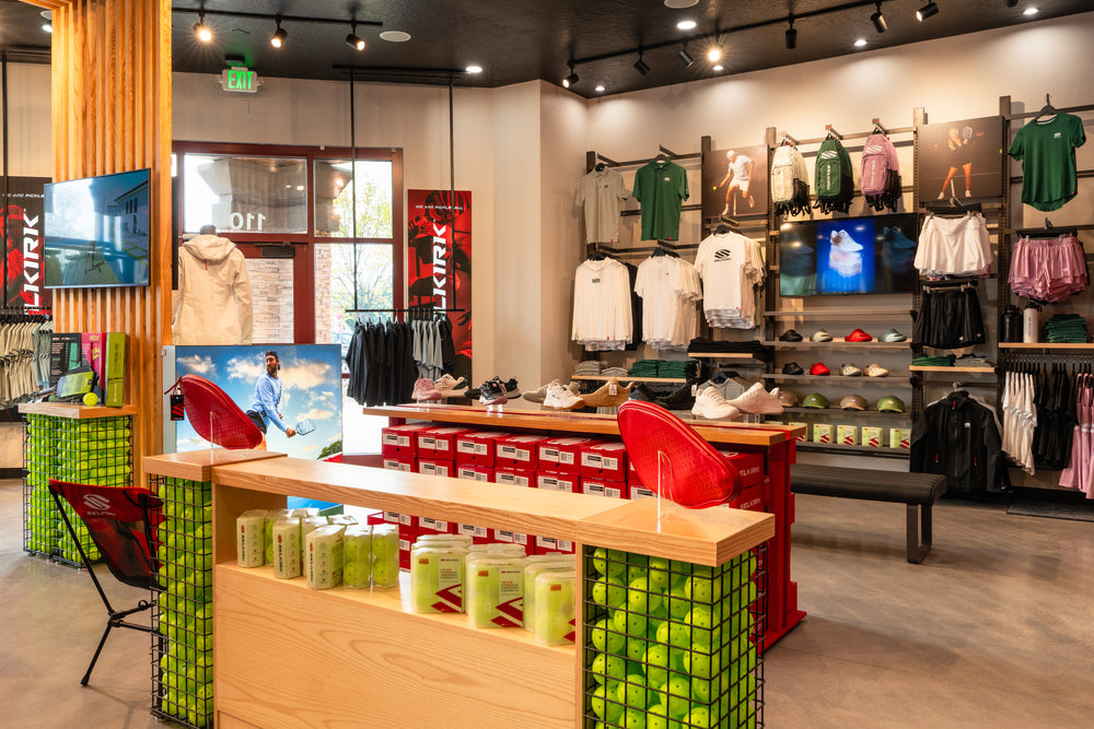 Pickleball shop interior with shoes, apparel, green ball racks and red boxes; EXIT sign and SELKIRK logo visible.