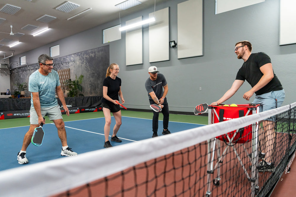 Four people practicing pickleball indoors; coach feeds balls from a red SELKIRK cart while three hold paddles by the net.