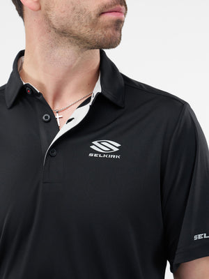 Male model wearing black polo with SELKIRK logo at chest, SEL on sleeve, and a visible cross necklace
