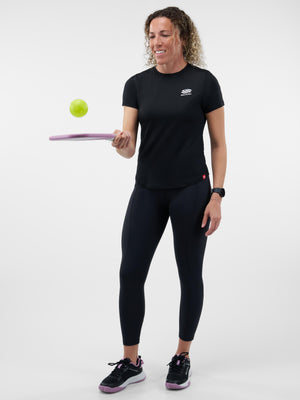 Woman in black athletic shirt and leggings balancing a green ball on a purple training paddle, smiling.
