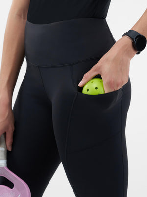 High-waist black leggings with a thigh pocket holding a green pickleball; wearer holds a paddle and wears a smartwatch.