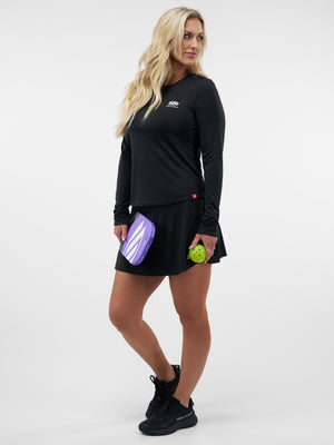 Woman in black athletic top and skirt holding purple pickleball paddle and green ball, wearing shoes labeled SELKIRK.