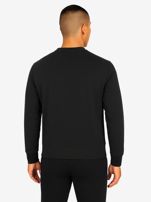 Back view of a man wearing a black crewneck sweatshirt and matching black pants, standing with hands relaxed at sides.