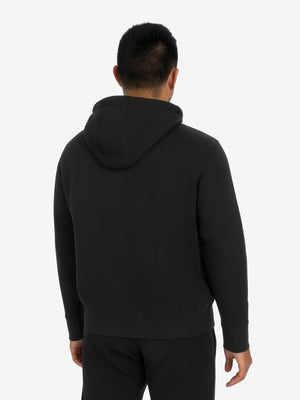 Rear view of a man wearing a black hoodie and black pants against a plain light background.