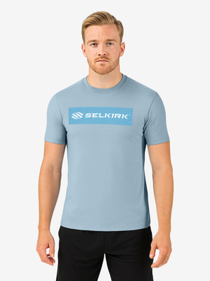 Man wearing light-blue short-sleeve Selkirk tee with SELKIRK wordmark centered in a darker-blue rectangular chest panel.