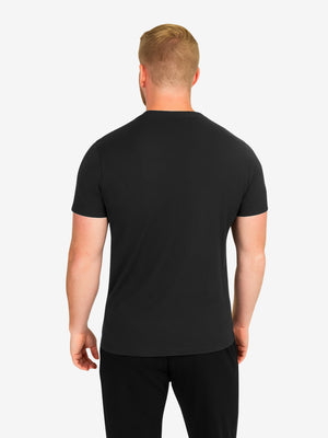 Back view of a man wearing a black crew-neck t-shirt and black pants on a light background.