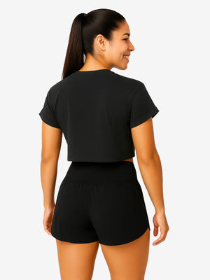 Back view of smiling woman with ponytail wearing a black cropped T-shirt and high-waist black athletic shorts.