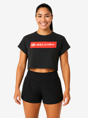 Woman wearing black crop T-shirt with red SELKIRK box logo, smiling, standing in black athletic shorts.
