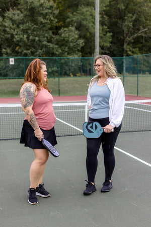 Two women on a tennis court, one wearing the AvaLee Crossover Tank in Arizona Sun, the other in a pink top.